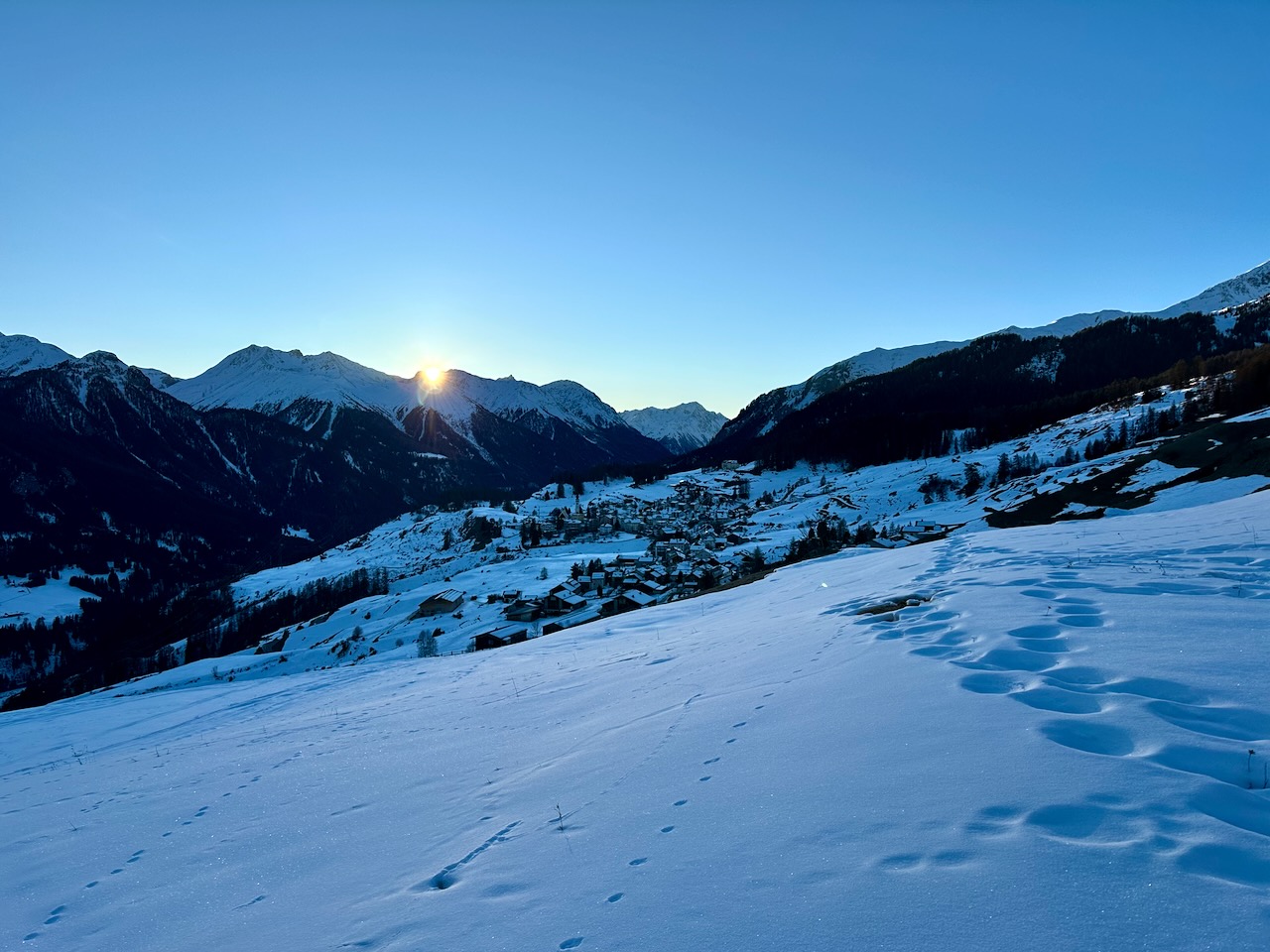 A snowy landscape in the Swiss Alps, with the sun just disappearing behind a mountain peak.